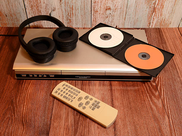 silver disc player with black headphones lying on it and an open plastic case with two compact discs against a wooden wall background, audio system with a remote control unit