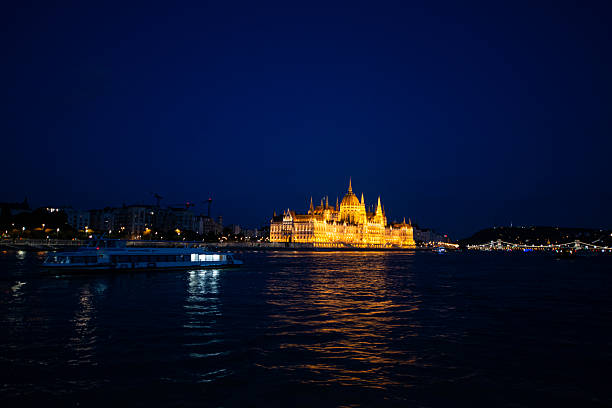 Night view of the Hungarian Parliament Building illuminated along the Danube River in Budapest, Hungary. The neo-Gothic landmark is reflected in the river waters, with the Chain Bridge visible nearby under a deep blue evening sky. The scene highlights one of Budapest’s most iconic architectural symbols and a UNESCO World Heritage site during nighttime.