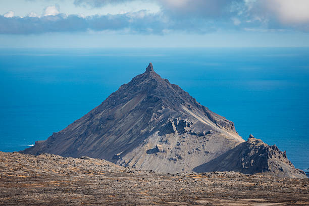 Iconic volcanic mountain peak rising from the rugged, rocky landscape of Snæfellsnes peninsula, with the vast Atlantic Ocean stretching to the horizon under a clear blue sky