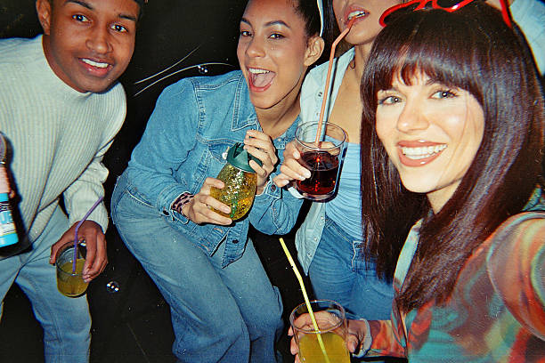 Group of young adult friends smiling and looking at the camera while enjoying drinks and dancing together at a lively party, captured with a vintage film aesthetic