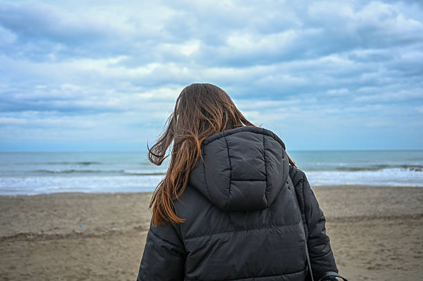 Young adult woman standing on a sandy beach, looking at the ocean under a cloudy sky