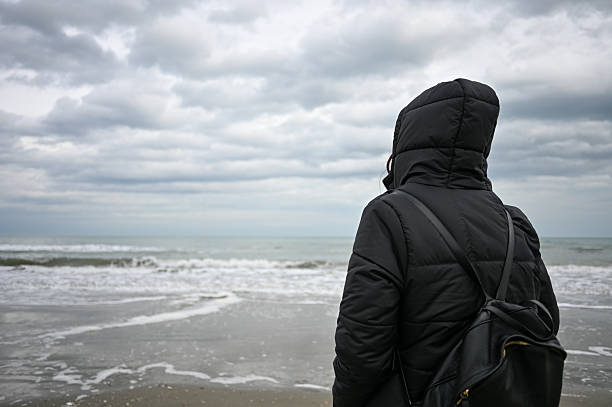 Woman in a black winter coat looking out at the cloudy ocean waves on a melancholic day