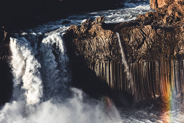 Aldeyjarfoss waterfall showcasing powerful water cascading over a dramatic wall of dark basalt columns, with a vibrant rainbow forming in the mist, highlighting Iceland's unique volcanic geology