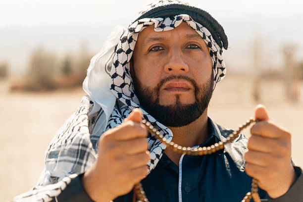 Close-up portrait of an Arab man holding a string of traditional beads toward the camera, wearing cultural clothing in a desert setting, expressing spirituality and identity.