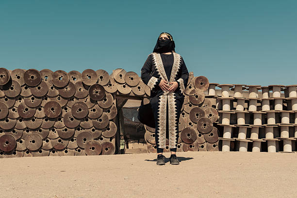 In the stillness of the desert, a young woman dressed in traditional Southwest Asian garb stands with quiet grace. The wooden shelter behind her adds texture to the scene filled with warmth and history.