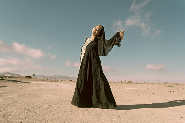 A joyful young Middle Eastern woman stands barefoot in the desert, smiling and enjoying the warmth of the sun and the freedom of the open space.