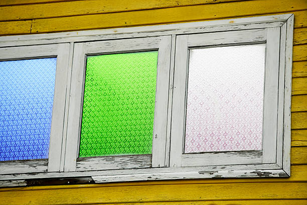 This image showcases a beautiful stained glass window with vibrant green, blue, and white hues, framed in a rustic white wood setting against a sunny yellow wall.