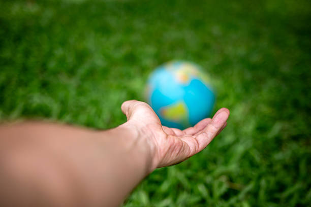 Human hand extended toward a blurred globe resting on green grass, using selective focus to emphasize the abstract concept of global responsibility, environmental fragility, and the future of the planet. Conceptual image representing sustainability, climate change awareness, environmental protection, and care for Earth, with ample copy space.