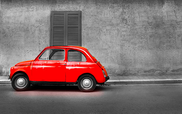 A timeless vintage Fiat 500 is parked against a textured brick wall, partially obscured by lush greenery and soft shadows. The black-and-white composition highlights the retro charm of the iconic car and its natural surroundings, creating a serene and nostalgic atmosphere