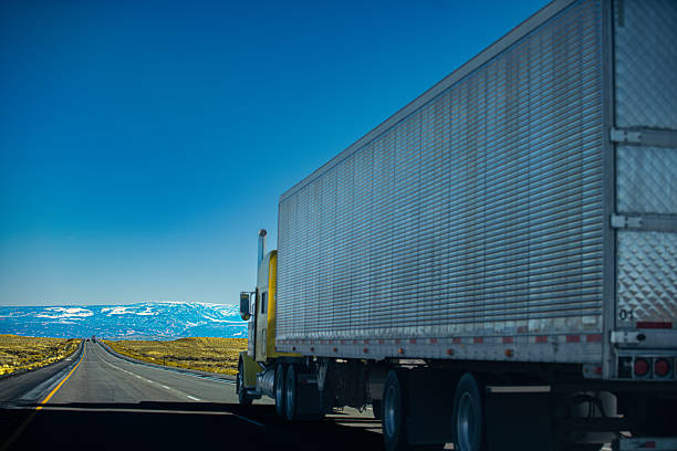A large white commercial truck travels along an empty highway with snow-capped mountains in the background. The road stretches through an open landscape with clear blue sky, representing long-distance transportation and logistics in a remote scenic environment.