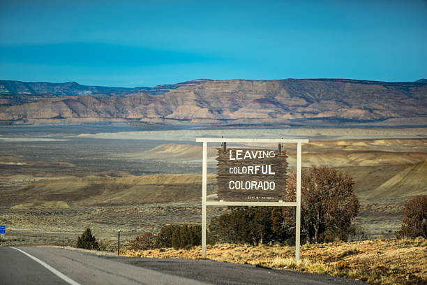 A roadside sign marks the boundary of Colorado, positioned against a dramatic desert landscape with rocky mountains and expansive plains. The sign reads 'LEAVING COLORFUL COLORADO', capturing the moment of geographic transition with a clear blue sky and rugged terrain in the background.