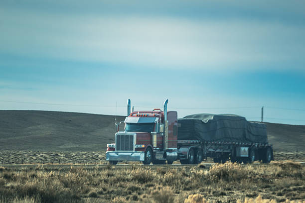 Large semi-truck transporting goods across a sparse grassland landscape with rolling hills and a pale blue sky. The truck appears to be a classic American long-haul semi with a red and white color scheme, carrying a dark-colored trailer loaded with cargo. The surrounding terrain is characterized by dry, sparse vegetation and an expansive, open terrain typical of western United States regions.