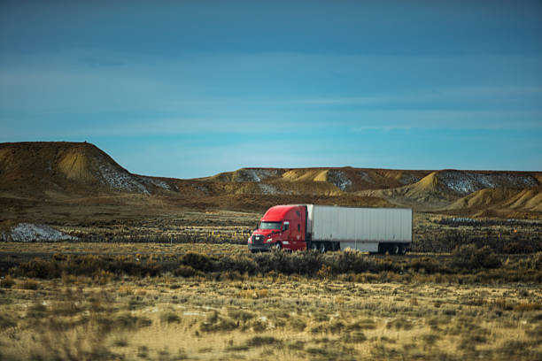 Red semi-truck with white trailer traveling through arid desert landscape with rocky hills and dry grasslands. Expansive blue sky provides backdrop to transportation scene showcasing long-distance trucking through remote wilderness area.