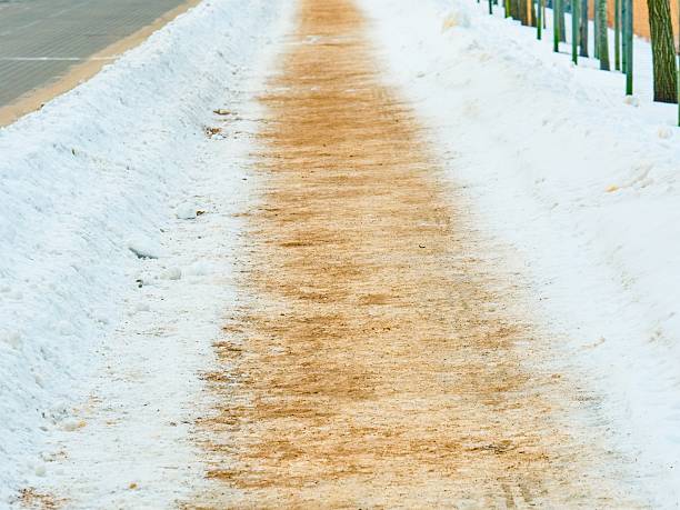 A pedestrian sidewalk covered in fresh snow with a clear path in the middle, treated with sand or grit for anti-slip protection during winter. The image conveys road maintenance and safety.