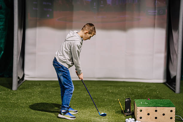 Young boy prepares to hit a golf ball indoors in a practice area featuring artificial turf and a screen showing a golf course landscape