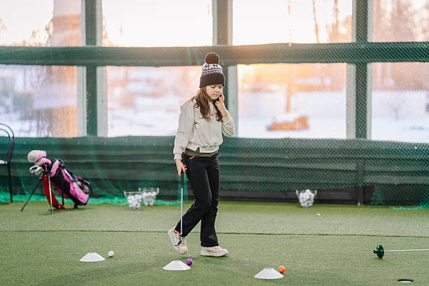 Young girl focuses on putting practice indoors at a golf training area, with artificial turf and golf equipment in the background