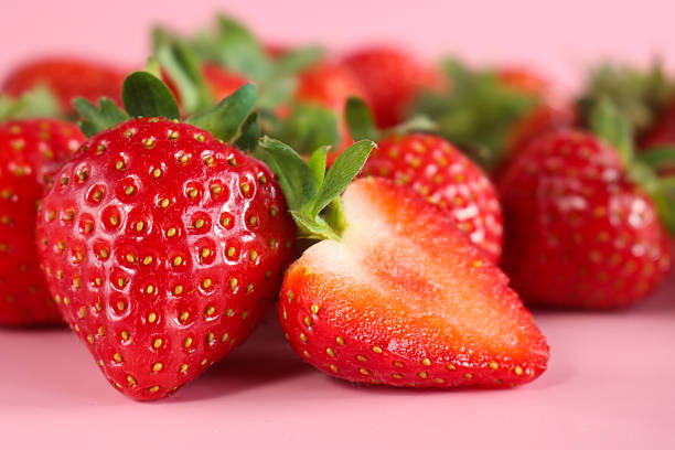 Close-up image of whole and halved fresh red strawberries with green leaves, cross section displaying inner juicy flesh, pink background,  focus on foreground, health and wellbeing concept