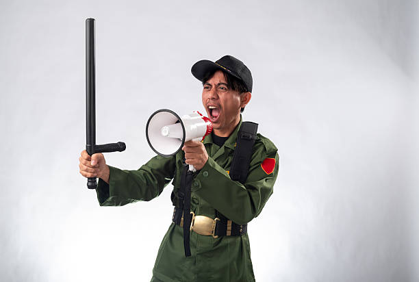 Asian male security officer in a green uniform and black cap, shouting through a megaphone while holding a baton, expressing authority and urgency, the plain white background