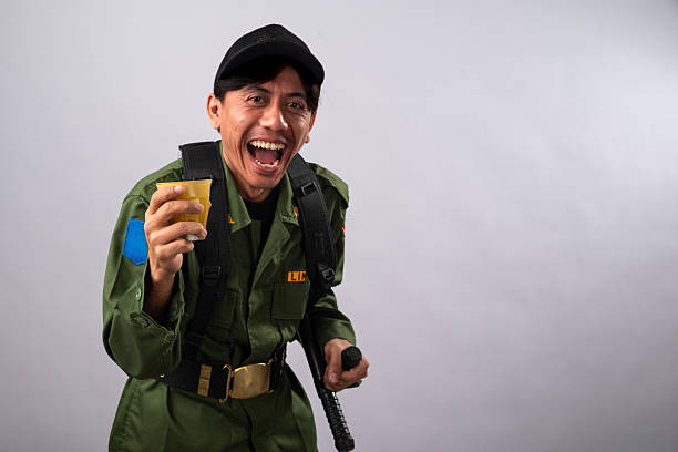 Asian male security officer in a green uniform and black cap, smiling widely while holding a drink in one hand and a baton in the other, the plain white background