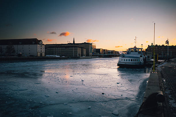 Ice covers Copenhagen Harbor in Denmark as a winter cold snap freezes sections of the city's waterways on February 3, 2026. A white passenger ferry sits frozen in the icy harbor with modern buildings visible along the waterfront during sunset. The unusual winter weather has created challenging conditions for maritime transport in Denmark's capital city.