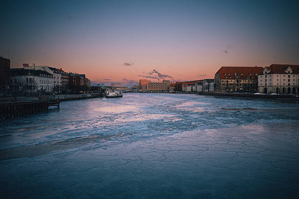 Ice covers Copenhagen's harbor waterway in Denmark during an unusual winter cold snap on February 3, 2026. The frozen harbor shows historic and modern buildings lining both sides of the waterway, with boats frozen in place along the ice-covered canal. The extreme winter weather has transformed the normally navigable Copenhagen waterways into a frozen landscape.