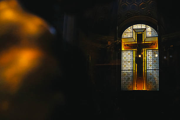 Cross silhouette in church interior against stained glass window. A blue sky with clouds and some vegetation can be seen behind. Concept for Christian religion, catholicism, prayer, hope and belief in God.