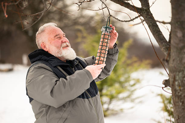 A senior man reaching up to secure a bird feeder on a tree branch, showcasing seasonal care for nature and wildlife during the cold winter months