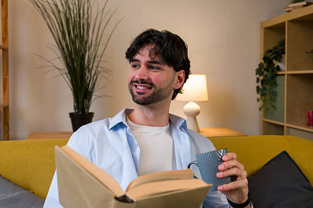 Young man smiling, holding coffee mug, and reading book on a comfortable sofa