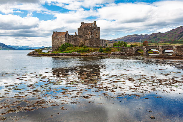 Eilean Donan Castle and stone bridge at low tide with seaweed in shallow water, set against Highland hills and dramatic clouds in Scotland