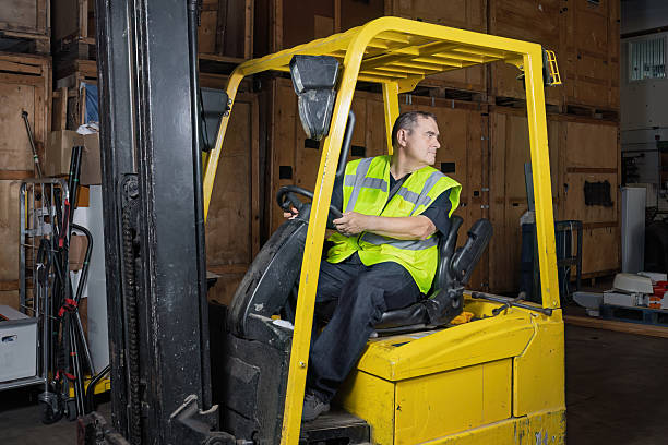 Man wearing high visibility vest reversing forklift truck inside warehouse while looking behind. Industrial workplace showing safe driving practice, materials handling, and health and safety awareness