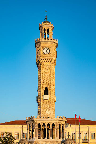 This wide-angle, low-view photograph captures the historic Antalya Clock Tower, built in 1901, standing tall against a blue sky. The Turkish flag waves proudly in the background, while tree branches in the foreground create a natural frame. The open-air shot highlights the harmony between the imposing structure and its natural surroundings. The image seamlessly blends cultural heritage with elements of nature, offering viewers a striking scene of both history and landscape.