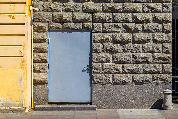 simple blue door stands embedded in a rough-hewn stone wall, flanked by a weathered yellow facade. The scene conveys a sense of quiet urban durability, with a security camera overhead adding a modern touch to the rustic setting.