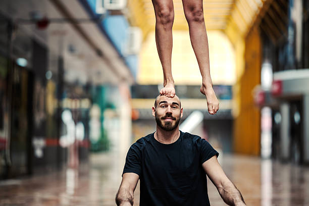 Young man with a beard concentrating while balancing female feet on his head, performing a complex acro yoga position indoors, demonstrating strength, trust, and partnership