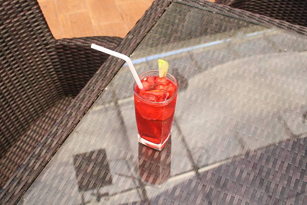 Vibrant red beverage served with ice cubes and a fresh lime garnish, resting on a reflective glass table surface, suggesting a relaxing outdoor moment