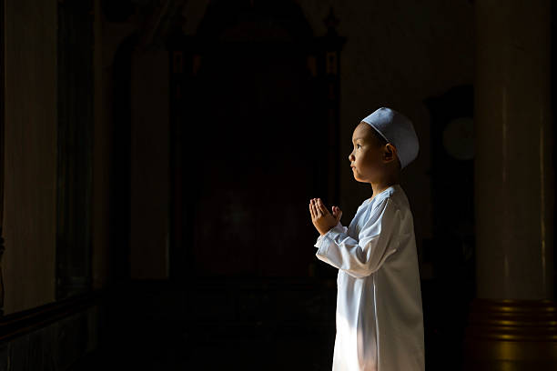 A young Asian Muslim boy standing and making a humble supplication (Du'a) inside a mosque with atmospheric lighting.