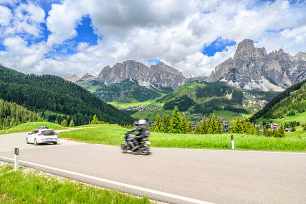View on Coravara and the Puez Group in Alta Badia Dolomites from the Passo Campolongo road during a springtime day with a motobike and car on the winding road, navigating a bend amidst lush green slopes and pine forests. In the midground, the village of Corvara is nestled in the valley. The majestic, rugged peaks of the Puez Group dominate the background under a partly cloudy sky. This landscape captures the beauty of the South Tyrol region during a bright springtime day, highlighting its appeal as a travel destination for outdoor activities like cycling.