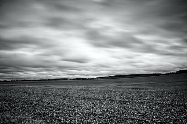 Moody black and white long exposure of an empty ploughed field under a dramatic sky with streaked, fast‑moving clouds. The minimalist rural scene in grey tones conveys solitude, quiet countryside atmosphere and fine art landscape aesthetics.