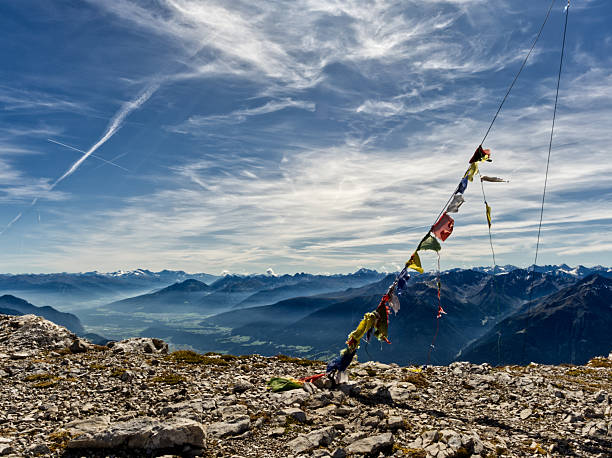 Colorful prayer flags flutter in the wind on a rocky alpine summit overlooking a vast mountain panorama and valley below under a blue sky with wispy clouds. The high altitude scene combines adventure, spirituality and the sense of freedom and achievement that comes with reaching the peak.