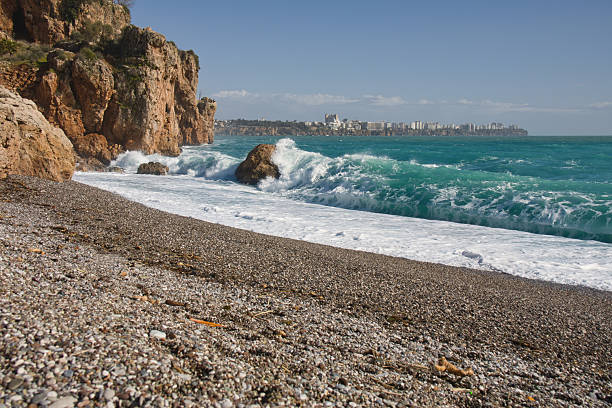 Powerful waves hitting the rocks at the start of Konyaalti cliffs in Antalya after a storm. Featuring turquoise waters, white sea foam, a pebble beach, and the city skyline in the background, capturing the raw energy of nature and urban life.