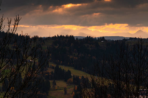 Distant mountain ridges beneath heavy storm clouds with fiery sunset glow, dark forested foreground and atmospheric depth in rural landscape