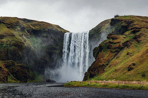 Skogafoss waterfall dramatically plunging over cliffs into a glacial river, surrounded by vibrant green hills on a gloomy day, showcasing Iceland's raw natural beauty and powerful landscapes