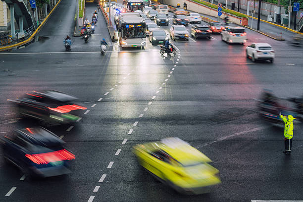 A busy Beijing intersection at dusk shows moving cars, buses, motorcycles, and a traffic marshal in bright yellow directing flow. Wet streets reflect lights, capturing urban energy, motion, and everyday city life.