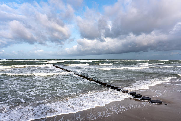 Dramatic Baltic Sea coastline near Ahrenshoop, Germany, featuring crashing waves and a long wooden breakwater stretching into the water beneath a heavy, cloudy sky.