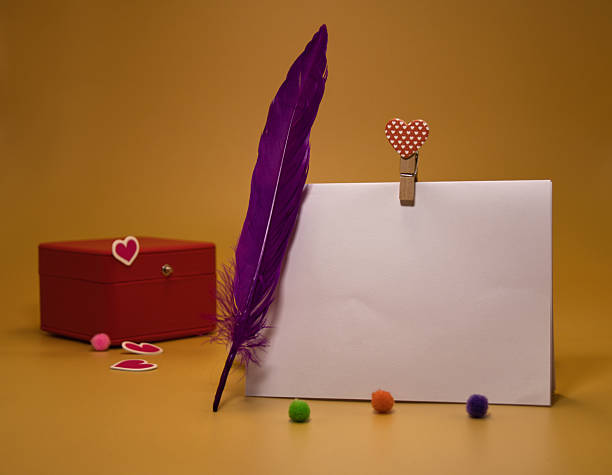 A blank white card placed horizontally on a gold-colored background. Includes a red gift box, a purple feather, a heart-shaped wooden clip, and colorful pom-poms. A romantic minimalist template with space to add text for Valentine's Day, love, or jewelry advertisements.