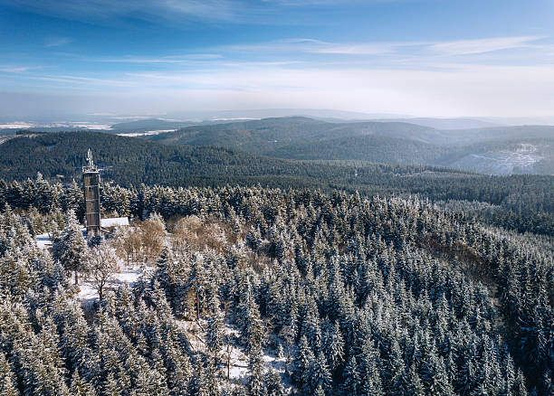 Aerial photograph of a snow-covered forest near Kickelhahn Mountain, Ilmenau, in Thuringia, Germany. Pine trees dusted with frost, a tall tower rising among the trees, expansive hills in the distance.