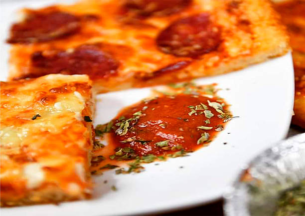 Close up of a pepperoni pizza slice served with tomato sauce and dried herbs on a white plate. Classic Italian comfort food concept ideal for restaurant menus and food advertising.
