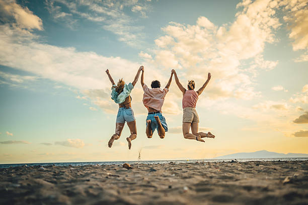 Happy young adult friends jumping and holding hands on a sandy beach under a beautiful sunset sky, celebrating freedom and summer vacation
