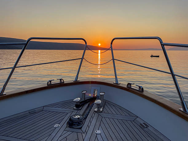 Wooden boat deck framing golden sunset over calm ocean waters with distant horizon and small boat silhouette in background.