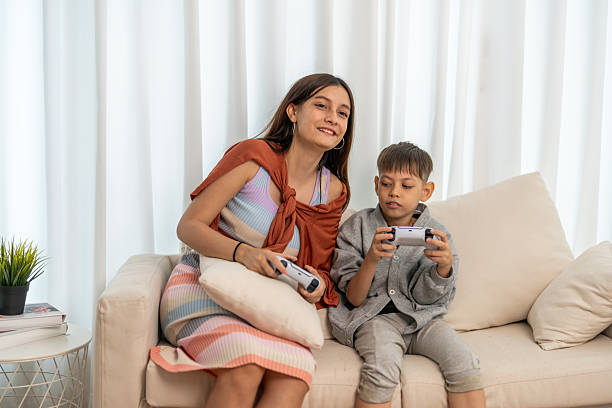 Two children sit side by side on a light-colored couch, each holding a game controller. The girl in the white dress appears very engaged, while the other girl looks at her with a slight smile.