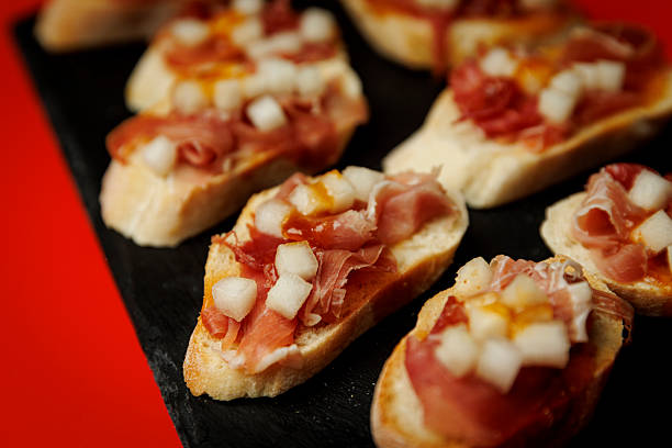Slices of bread topped with prosciutto, diced pear, and sauce are arranged on a black slate surface. The close-up emphasizes texture and color in the appetizer.